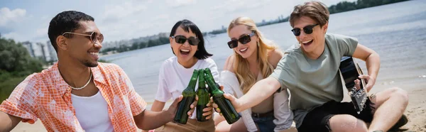 Cheerful interracial friends in sunglasses clinking bottles during beer party, banner — Stock Photo