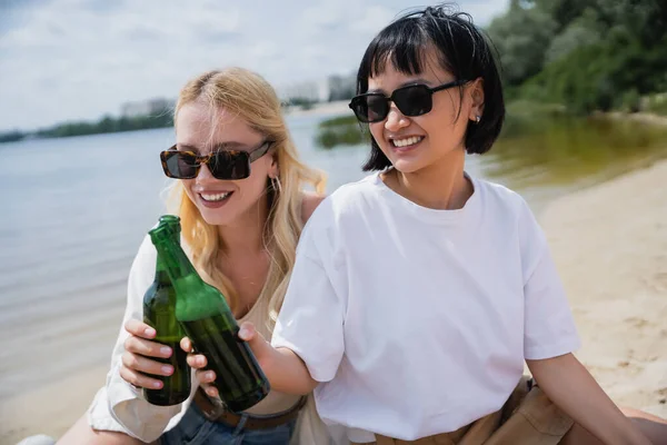 Smiling multiethnic friends in sunglasses drinking beer bottles on beach — Stock Photo