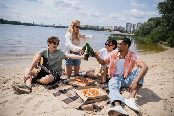 Excited multiethnic friends in sunglasses clinking beer bottles during picnic on beach — Fotografia de Stock