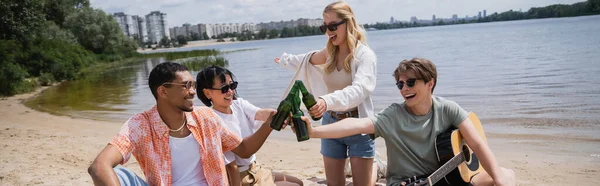 Happy woman pointing with hand near multiethnic friends clinking bottles of beer, banner - foto de stock