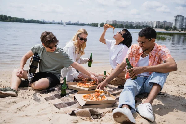Young multiethnic friends eating delicious pizza and drinking beer on beach — Fotografia de Stock