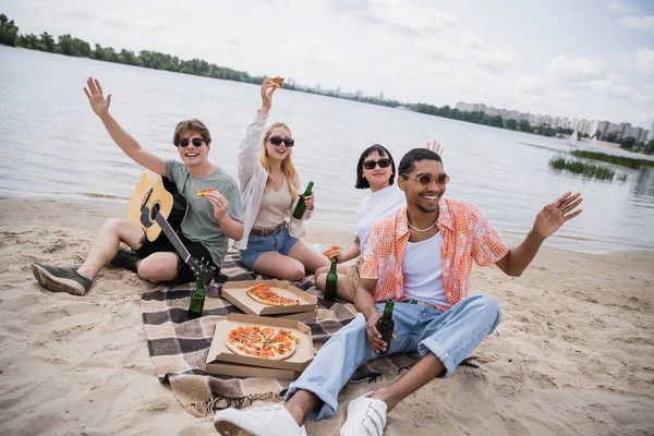 Cheerful multiethnic friends waving hands during picnic on riverside - foto de stock