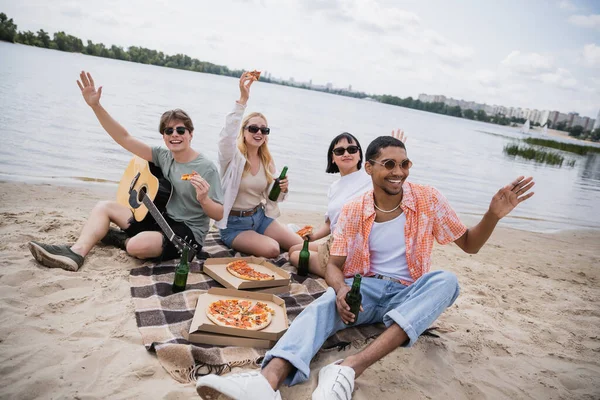 Cheerful multiethnic friends waving hands during picnic on riverside — Fotografia de Stock