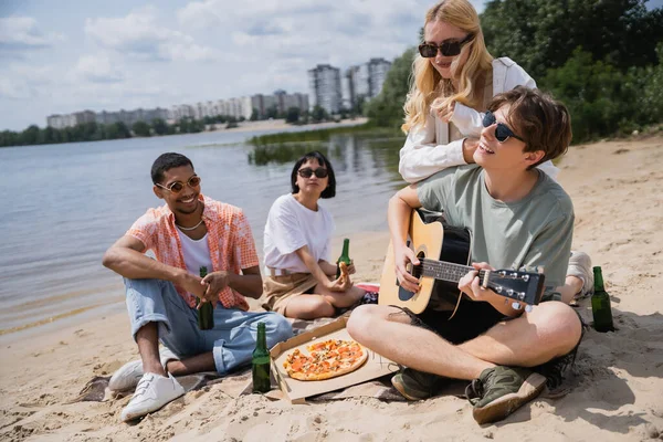 Man in sunglasses playing guitar during beach party with interracial friends — Stockfoto