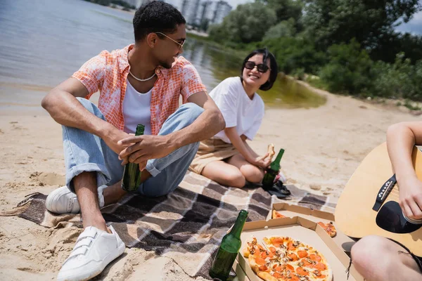 Interracial friends looking at each other near man playing guitar during picnic — Stock Photo