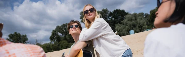 Smiling man in sunglasses playing guitar near blonde woman and interracial friends, banner - foto de stock