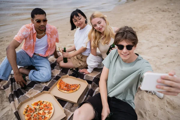 Man in sunglasses taking selfie with friends during picnic on riverside — Photo de stock