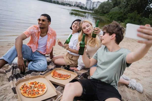 Happy man taking selfie on blurred smartphone during beach picnic with interracial friends — Stock Photo