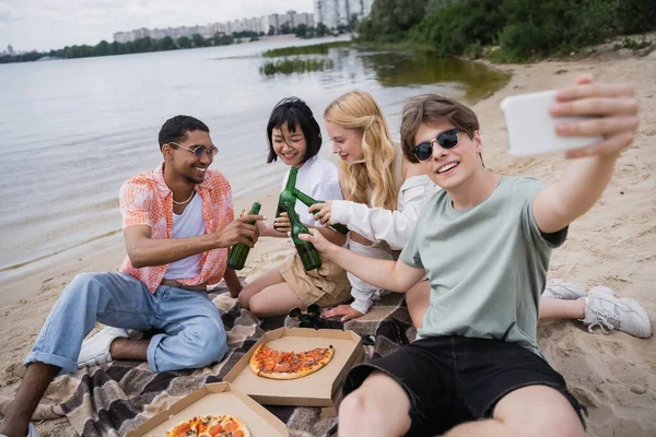 Smiling man taking selfie with multiethnic friends clinking beer bottles near pizza — Fotografia de Stock