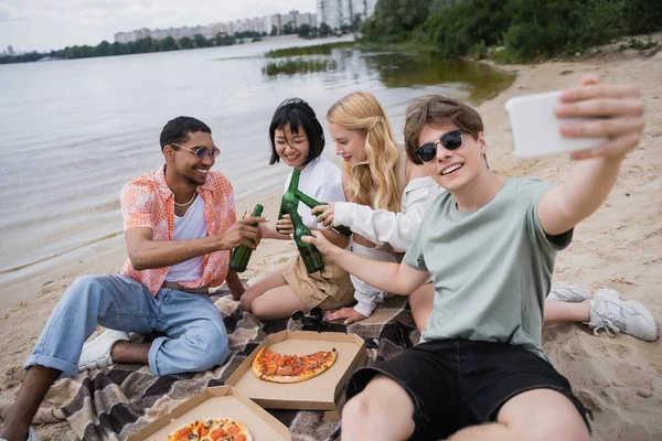 Smiling man taking selfie with multiethnic friends clinking beer bottles near pizza — Stock Photo