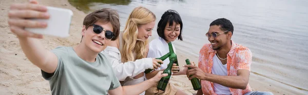 Man in sunglasses taking selfie on blurred smartphone during beer party with interracial friends, banner - foto de stock