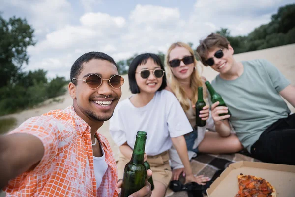 Молодые и счастливые межрасовые друзья с пивом, смотрящие в камеру возле пиццы на пляже — стоковое фото