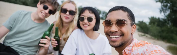 Cheerful african american man smiling at camera near blurred friends with beer, banner — Stockfoto