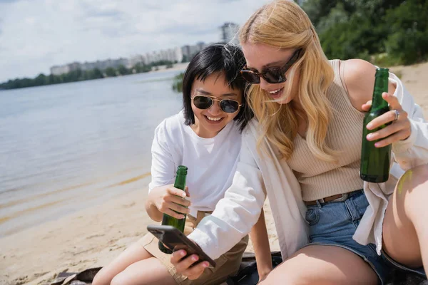 Cheerful interracial women in sunglasses and with beer bottles looking at smartphone on beach — Stock Photo