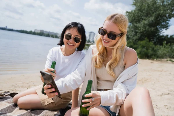Blonde woman in sunglasses showing smartphone to asian friend with bottle of beer — Foto stock
