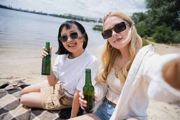 Asian woman sticking out tongue while drinking beer with blonde friend on beach — Stock Photo