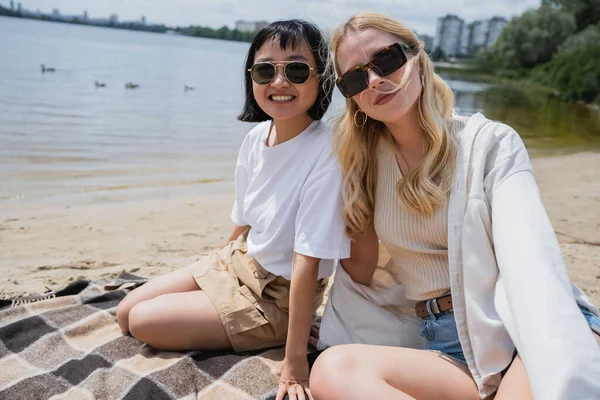 Young interracial friends in sunglasses sitting on blanket near river and looking at camera — Stock Photo