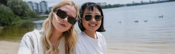 Cheerful interracial women in sunglasses looking at camera near river, banner — Stock Photo