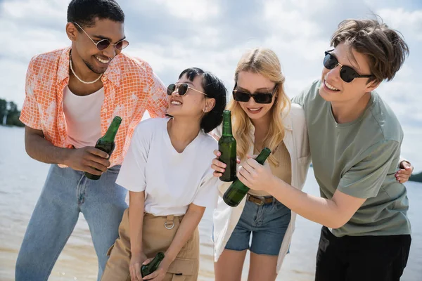 Joyful multiethnic friends holding beer bottles on beach — Stock Photo