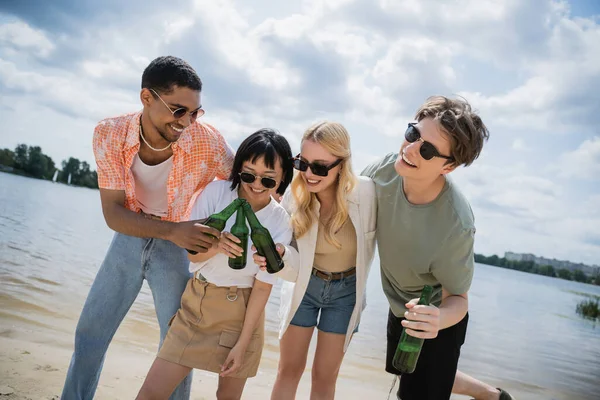 Low angle view of multicultural friends in sunglasses clinking refreshing beer on beach — Stock Photo