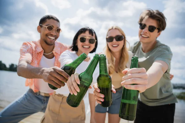 Blurred interracial friends in sunglasses clinking bottles during beer party — Stock Photo