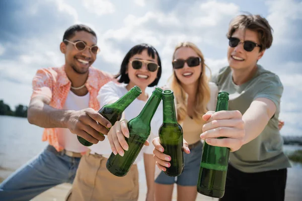 Interracial friends in sunglasses clinking beer bottles on blurred background — Stock Photo