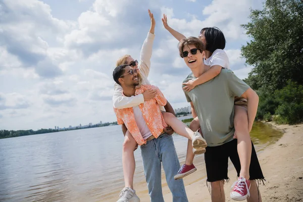 Cheerful women giving high five while interracial men piggybacking them near river — Stock Photo
