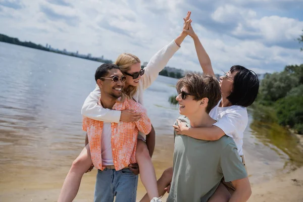 Interracial men piggybacking women giving high five on riverside — Stock Photo