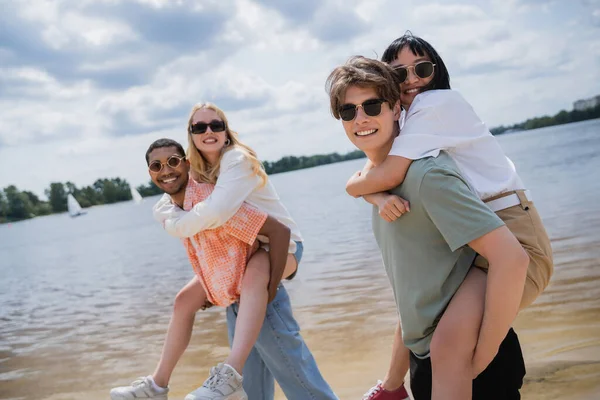 Young multicultural men in sunglasses looking at camera while piggybacking women near river — Stock Photo