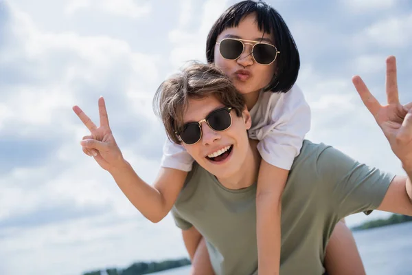 Man in sunglasses piggybacking asian girlfriend pouting lips and showing victory gesture — Stock Photo