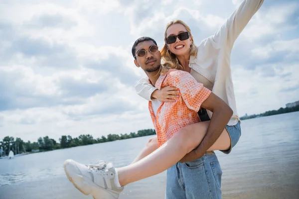 Young african american man piggybacking girlfriend in sunglasses near river — Stock Photo