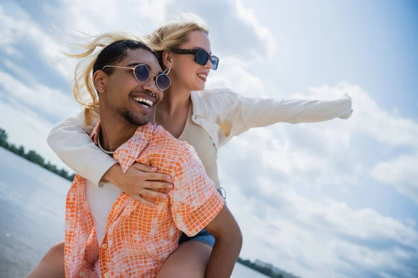 Happy african american man in sunglasses piggybacking excited girlfriend on summer day - foto de stock
