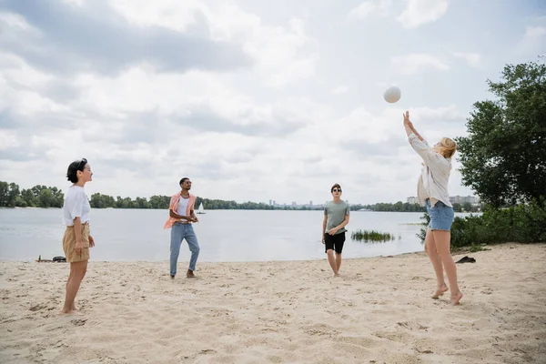 Young woman passing ball to multiethnic friends playing beach volleyball on riverside — Stock Photo