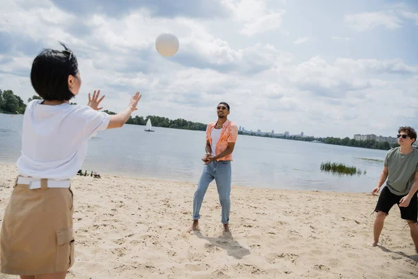 Young multiethnic friends playing volleyball on sand beach near river — Stock Photo