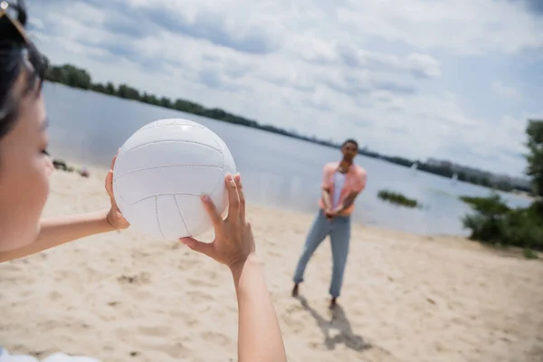 Asian woman holding ball while playing on beach with blurred african american man — Stock Photo
