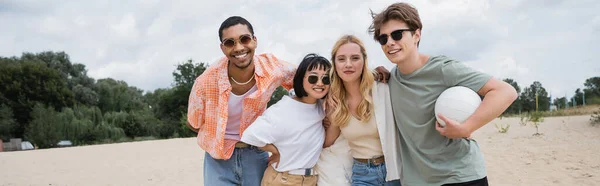 Young and happy interracial friends with ball smiling at camera on beach, banner — Stockfoto