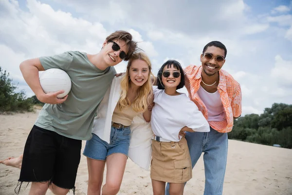 Cheerful multiethnic friends in sunglasses smiling at camera on beach — Stock Photo