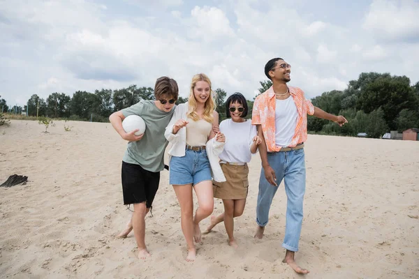 Возбужденные мультикультурные друзья прогуливаясь по песчаному пляжу с мячом — стоковое фото