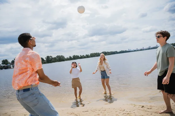 Joyful multiethnic friends playing beach volleyball near river on summer day - foto de stock