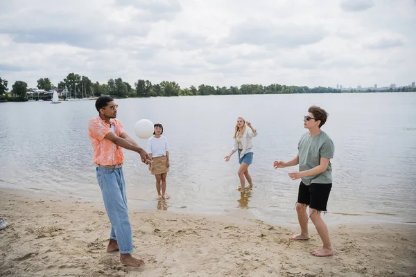 Interracial men playing beach volleyball near women standing in water — Stock Photo