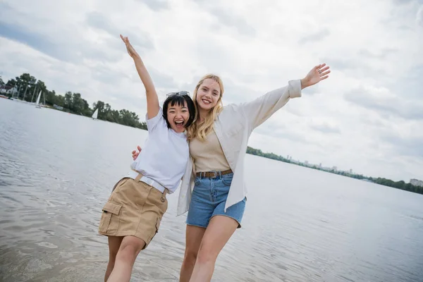 Excited interracial friends hugging and posing with raised hands near river — Fotografia de Stock