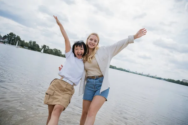 Excited interracial friends hugging and posing with raised hands near river — Stock Photo