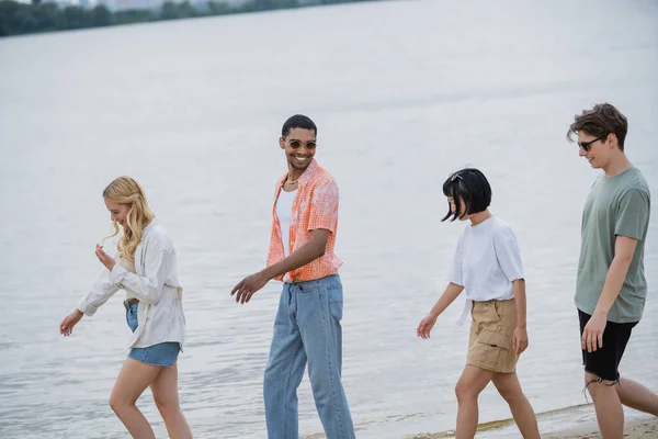 Cheerful african american man in sunglasses walking with interracial friends near river — Stock Photo