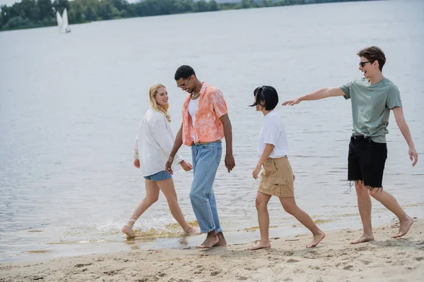 Young man in sunglasses pointing with hand near multiethnic friends walking on beach - foto de stock