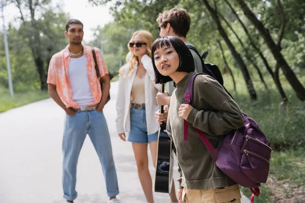 Young asian woman with backpack looking away near multiethnic friends on blurred background — Photo de stock