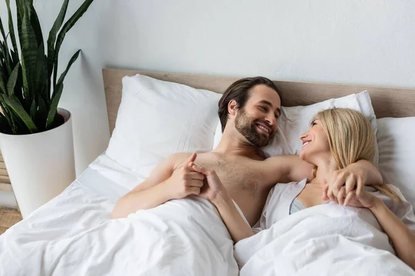 Happy lovers holding hands and smiling at each other on bed in morning — Photo de stock