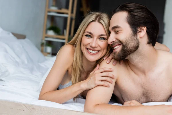 Happy blonde woman smiling at camera and hugging shirtless man in bed - foto de stock