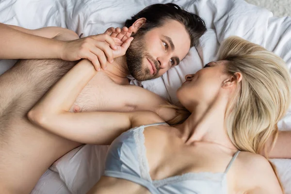 Вид сверху на брюнета, держащего за руку сексуальную женщину, глядя друг на друга в постели — стоковое фото