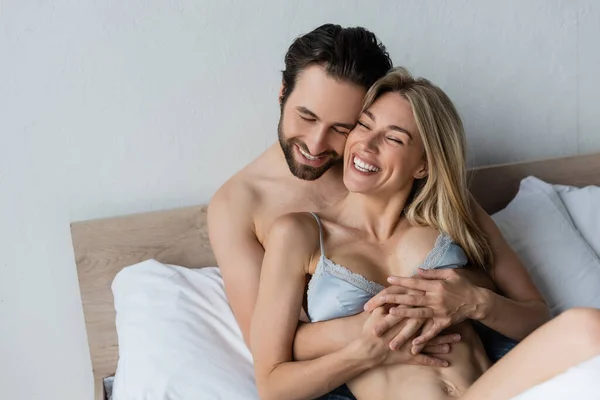 Улыбающийся бородатый мужчина обнимает счастливую женщину в сексуальном белье в спальне — стоковое фото
