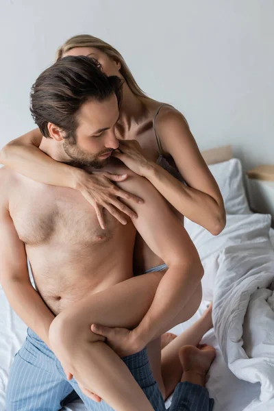 Passionate woman hugging shirtless man in pajama pants in bedroom — Stock Photo